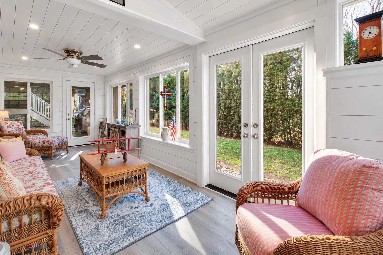 Bright sunroom with white paneled walls, large windows, wicker seating and a glass-paneled double door.