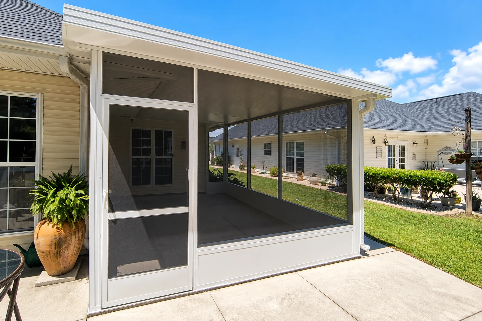 A white patio enclosure with dark screens and a sliding screen door stands beside a house on a concrete patio.