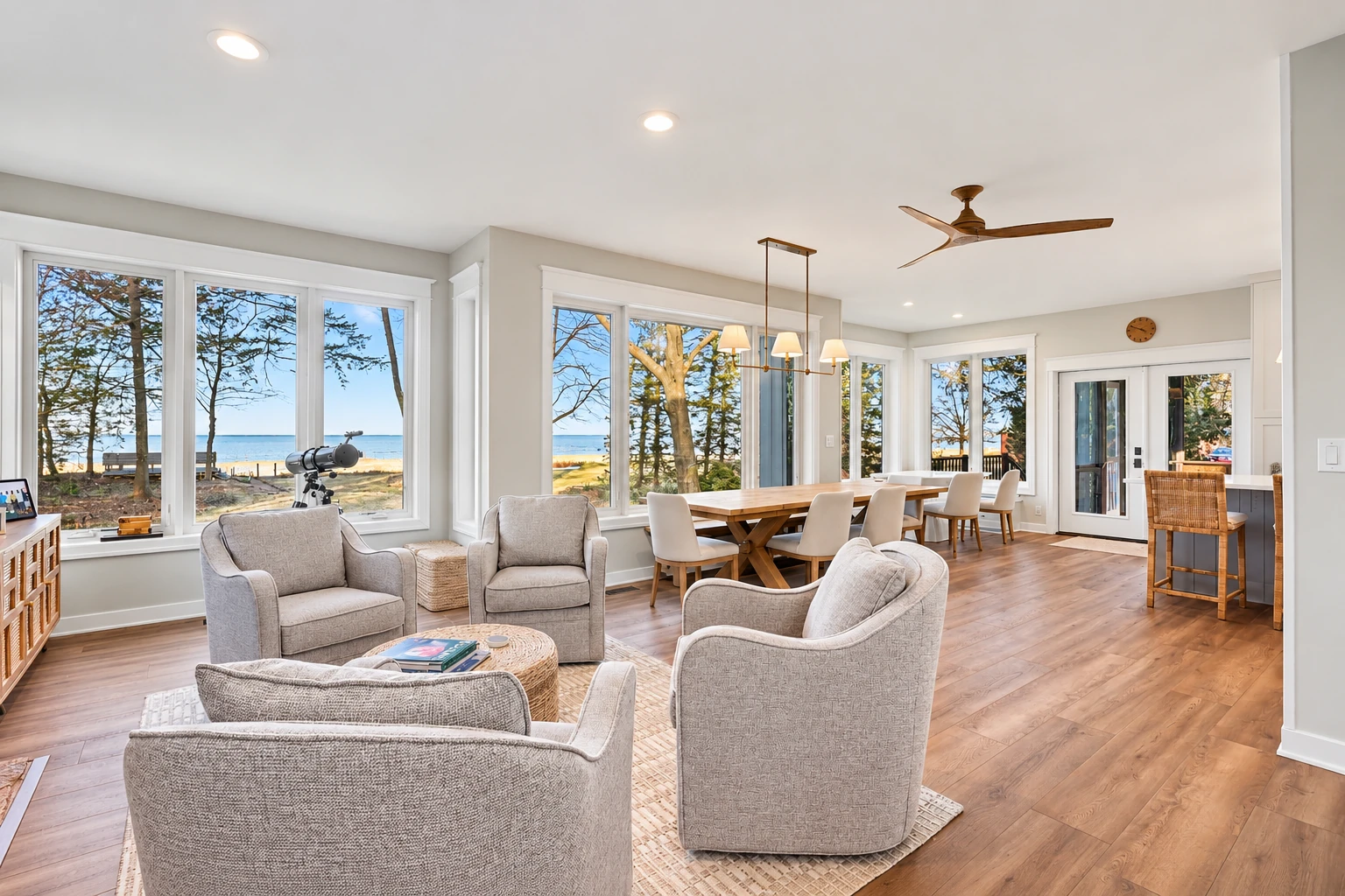 Bright sunroom addition with gray armchairs, a dining table, and large windows overlooking a lake.