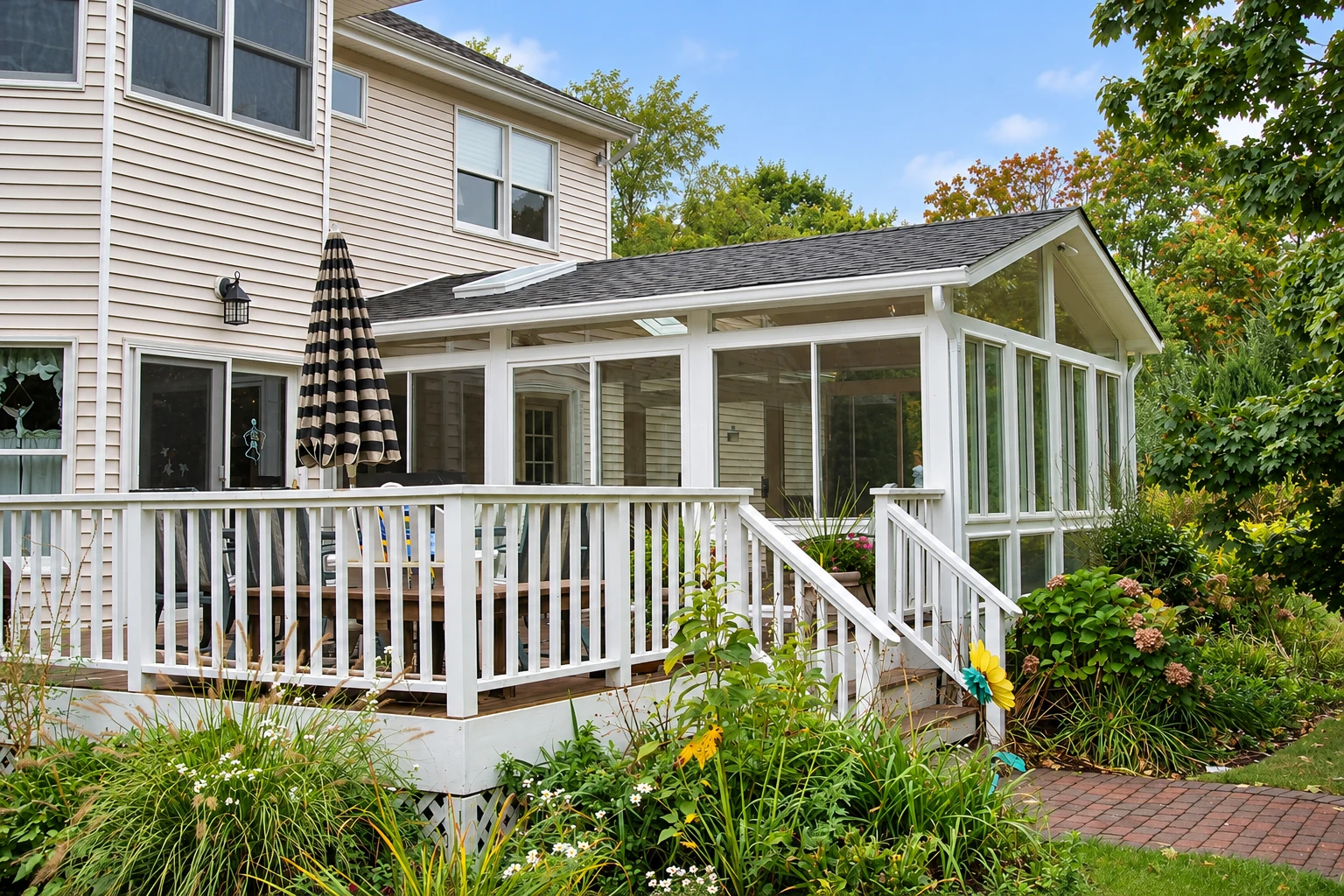 A white-framed sunroom addition with a peaked glass end wall extends from a two-story house beside a deck and garden.