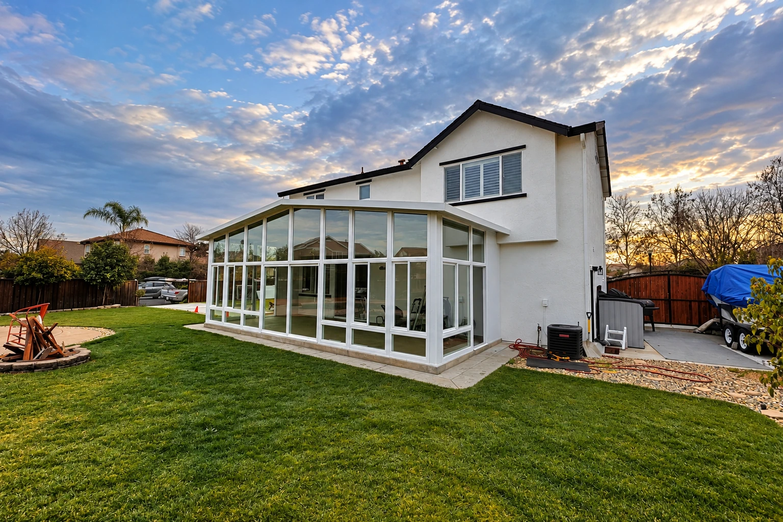 A white-framed glass sunroom addition extends from a two-story house into a green backyard beneath a cloudy sunset sky.