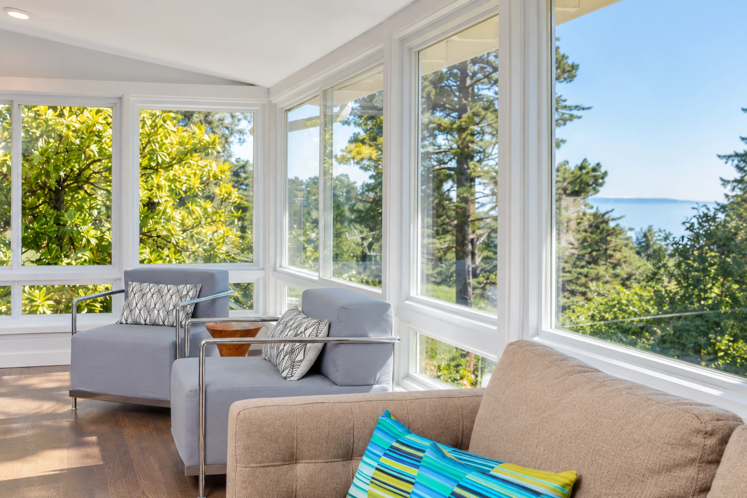 Bright sunroom with wraparound windows, two gray armchairs and a beige sofa overlooking trees and water.