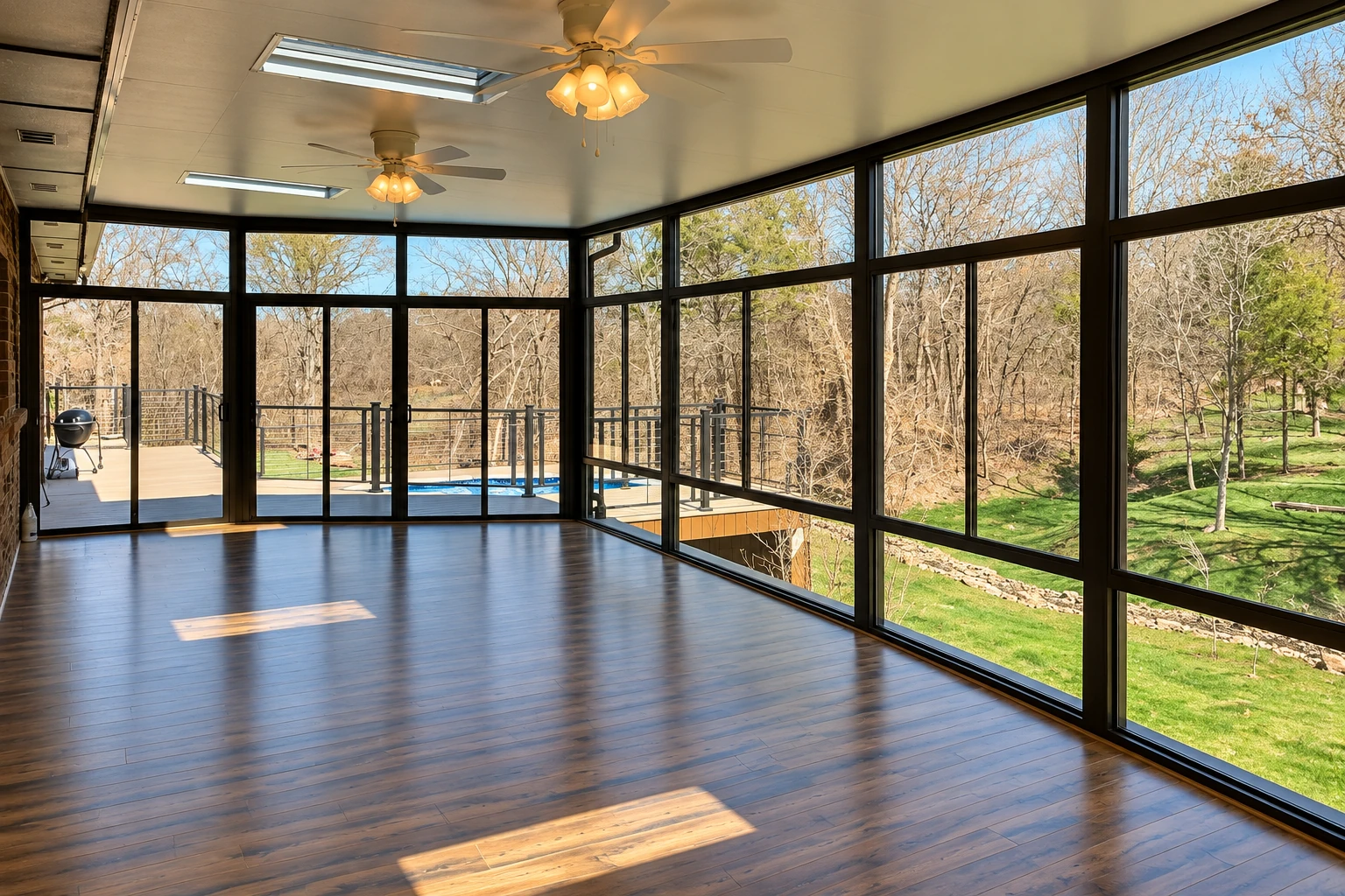 An empty remodeled sunroom has dark-framed windows, wood flooring, ceiling fans, and views of trees and a pool.