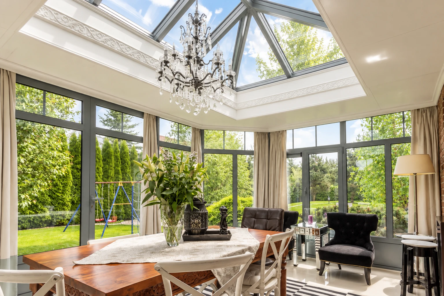 Sunroom with a glass gabled ceiling, tall windows, a chandelier and dining furniture overlooking a garden.